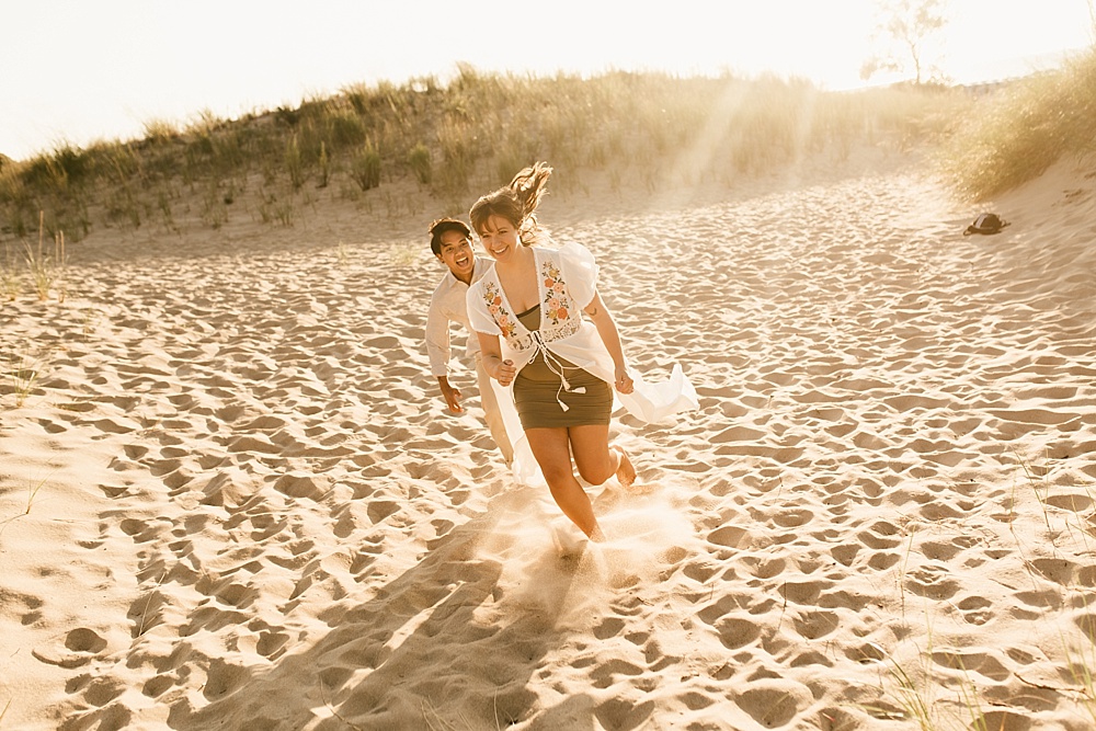 couple running down sand dune at warren dunes van life engagement shoot