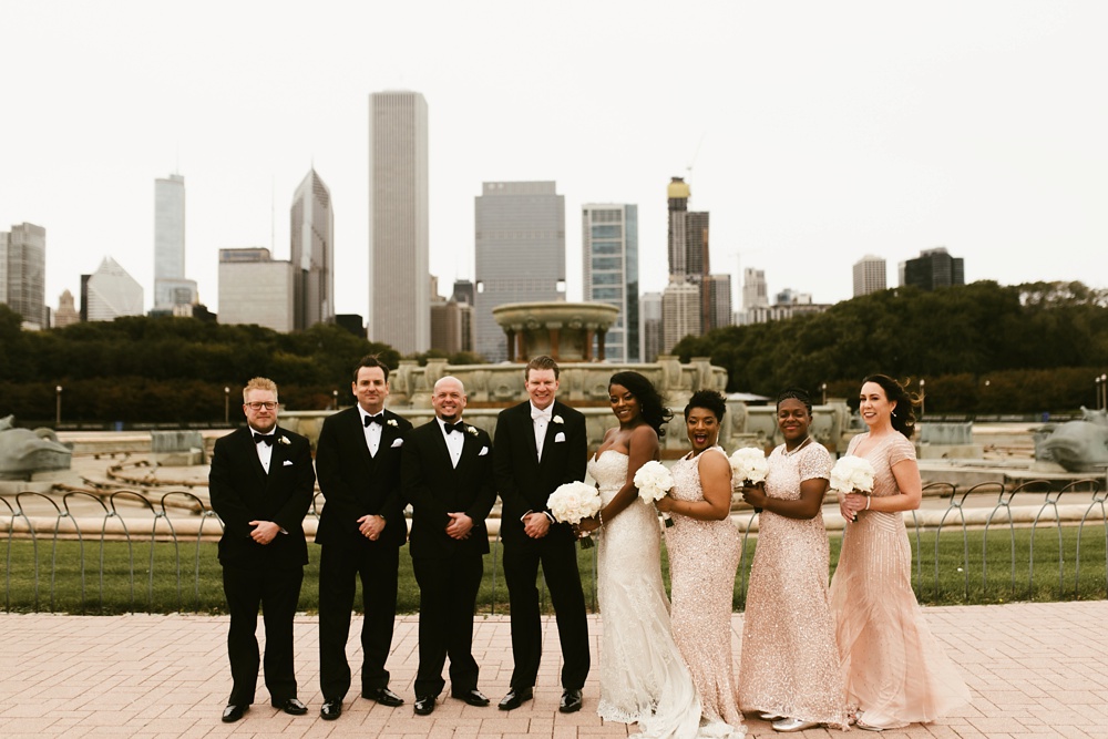 bridesmaids in rose dresses and groomsmen in black tuxes with bride and groom at buckingham fountain chicago