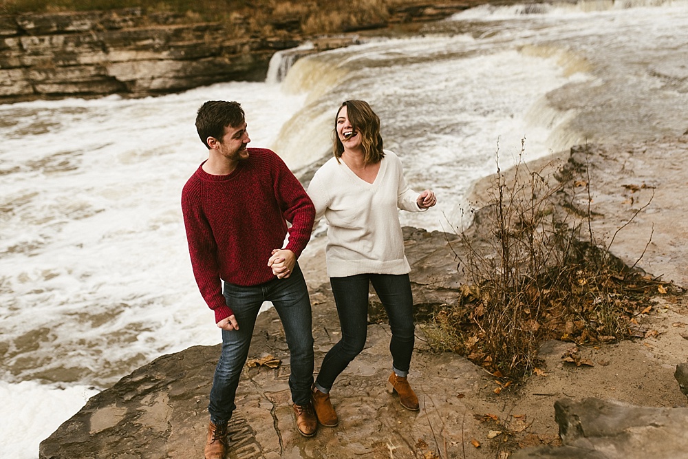 couple laughing and running next to falls at cataract falls fall engagement session