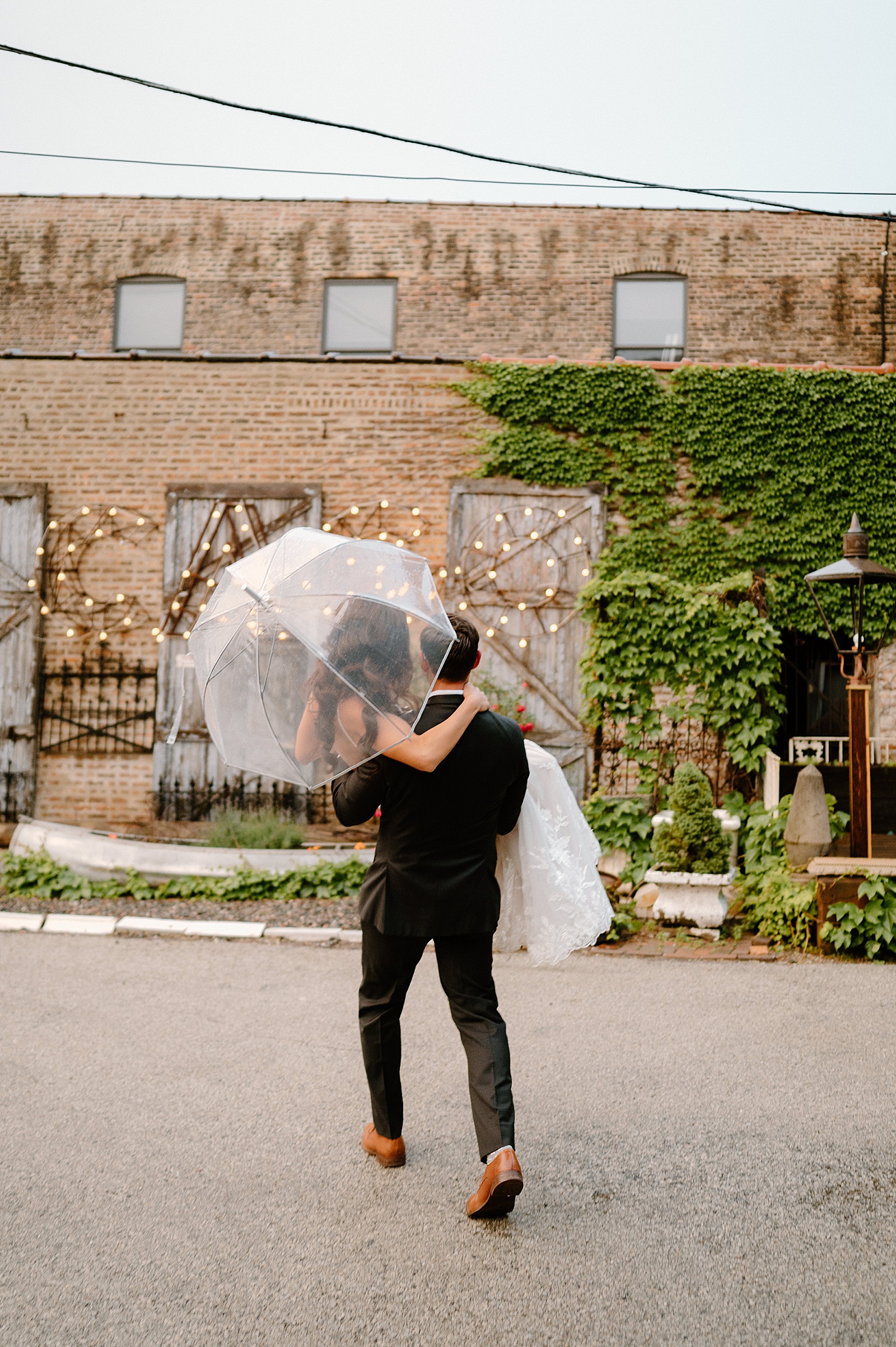 Groom carrying bride with clear umbrella in a courtyard for their rainy wedding day 