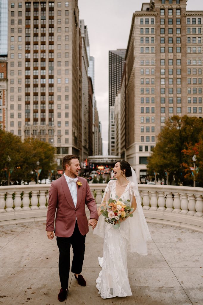 Bride in boho dress and groom in pink suit walking with Chicago in the background.