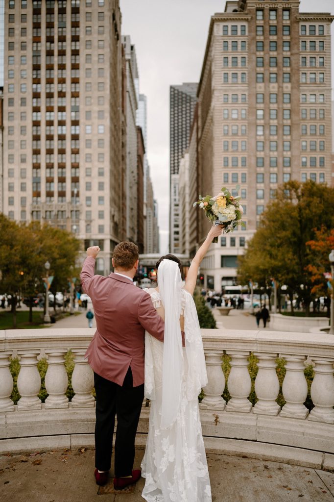 Bride and groom on a balcony overlooking downtown Chicago, throwing their hands in the air in celebration.