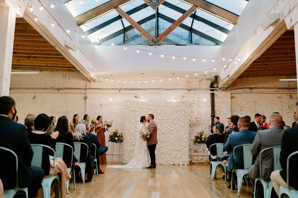 An indoor wedding ceremony with a floral wall installation and twinkle lights overhead. 