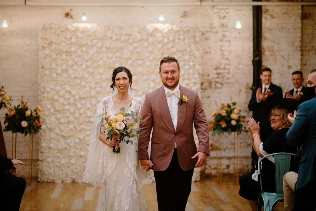 Bride and groom walking back down the aisle with floral installation on the wall behind them at The Joinery wedding venue. 