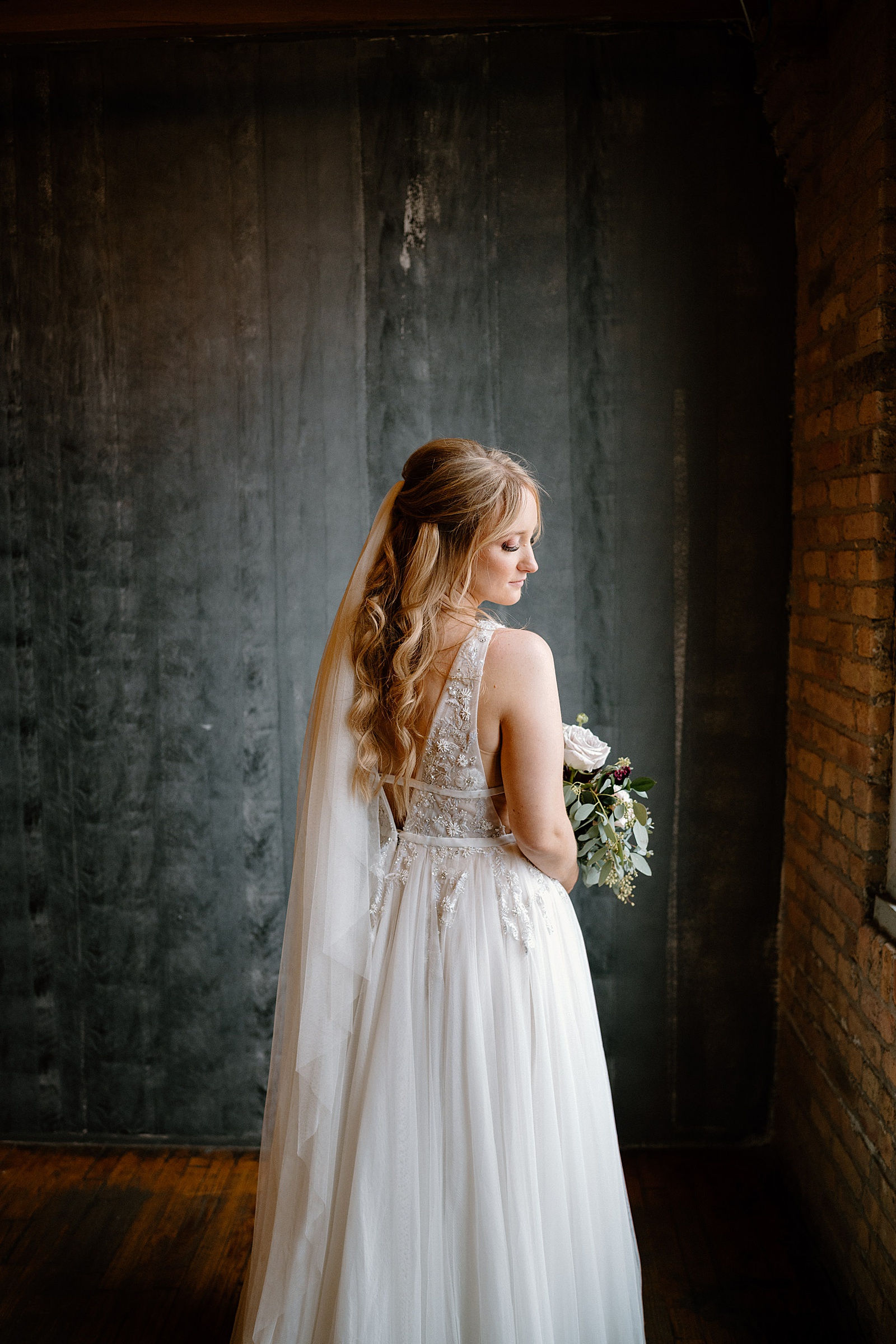 Bride in lace long gown in front of a window holding flowers