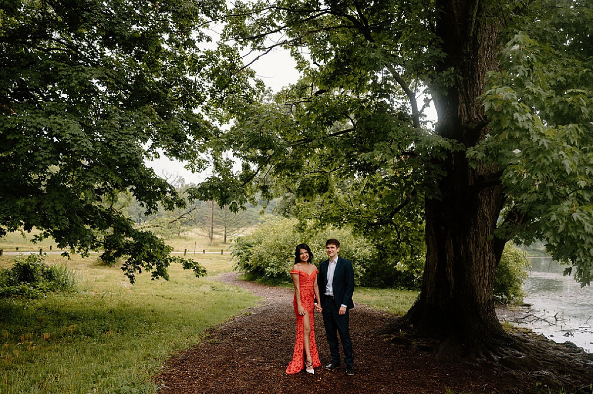 woman in red evening gown and man in black suit pose under large tree during mortem arboretum engagement session