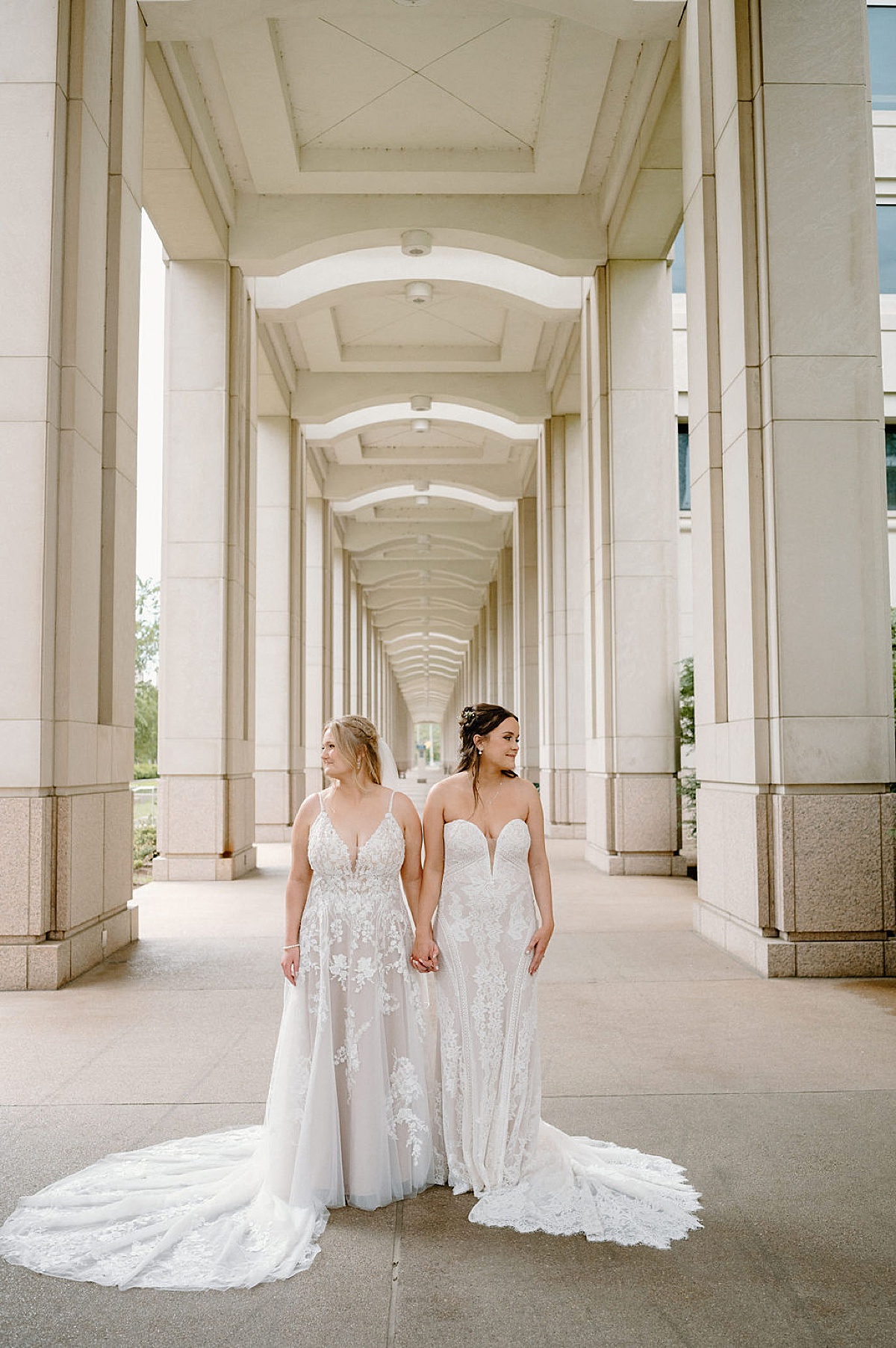 Two women in bridal gowns pose hand in hand between elegant columned walkway at ceremony by midwest wedding photographer