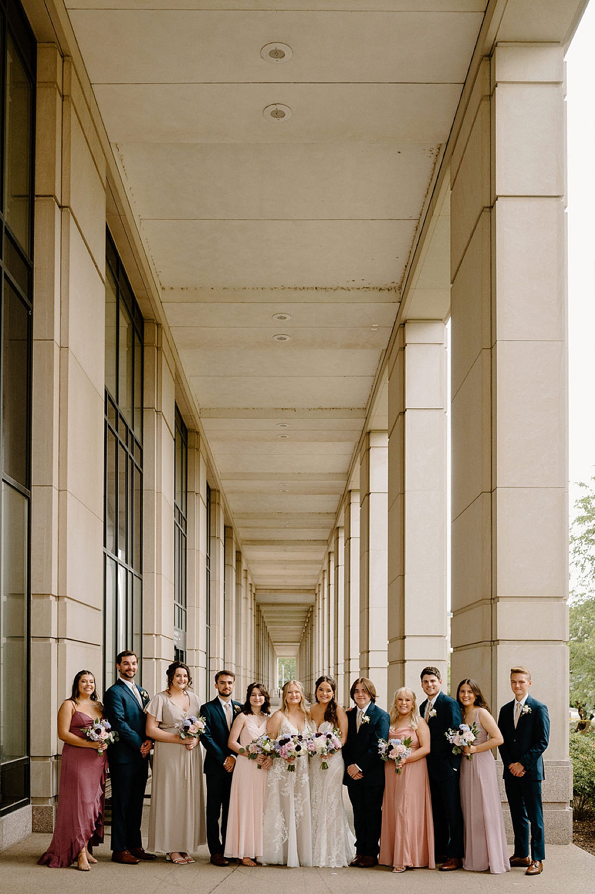 bridal party in soft pastel gowns and navy blue suits pose with two brides at ceremony shot by midwest wedding photographer