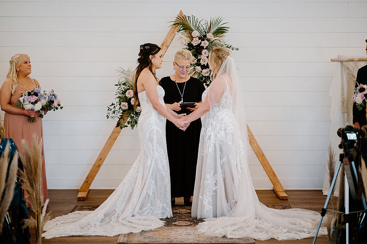 Two brides hold hands in front of boho floral arrangement during wedding ceremony shot by Indigo Lace Collective