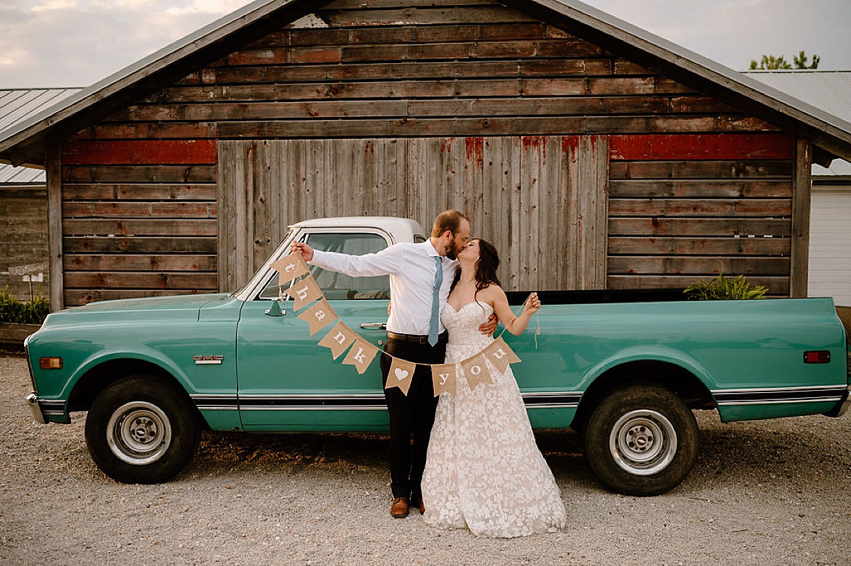 bride and groom kiss with thank you sign beside teal retro truck after rustic summer wedding at Heritage Prairie Farm