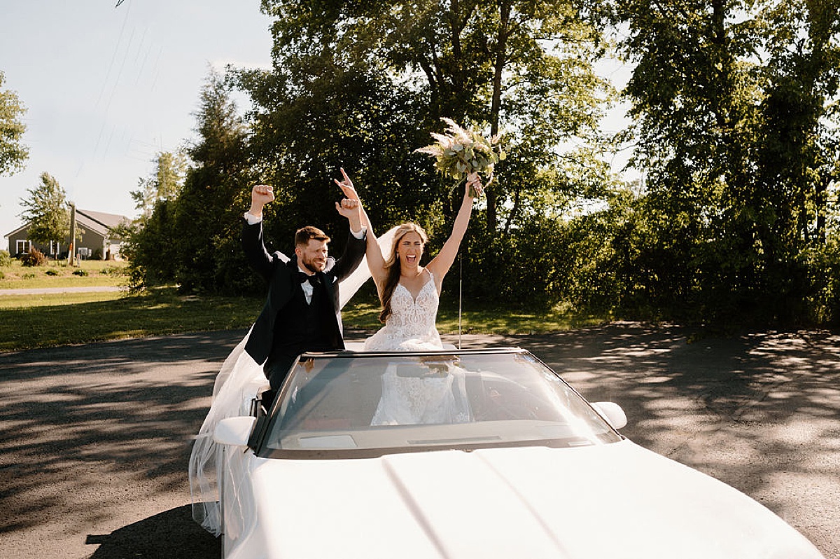 bride and groom sit in back of convertible and throw their hands in the air after good vibes wedding shot by indigo lace collective
