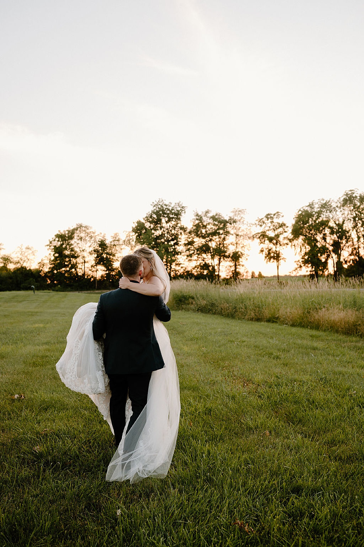 groom carries bride across a green midwest field during golden hour after wedding shot by indigo lace collective
