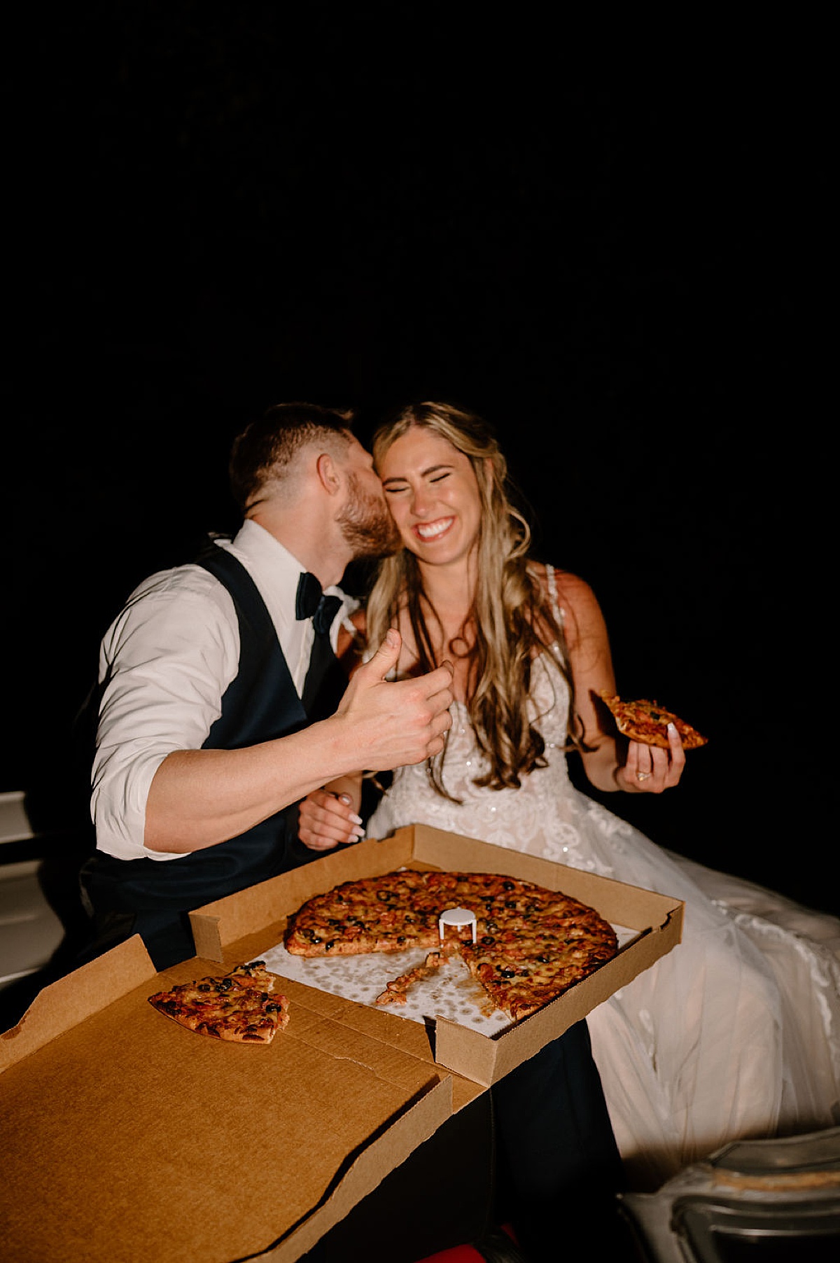 groom kisses bride on the cheek while the newlywed couple share a pizza in the back of a white convertible. shot by indigo lace collective