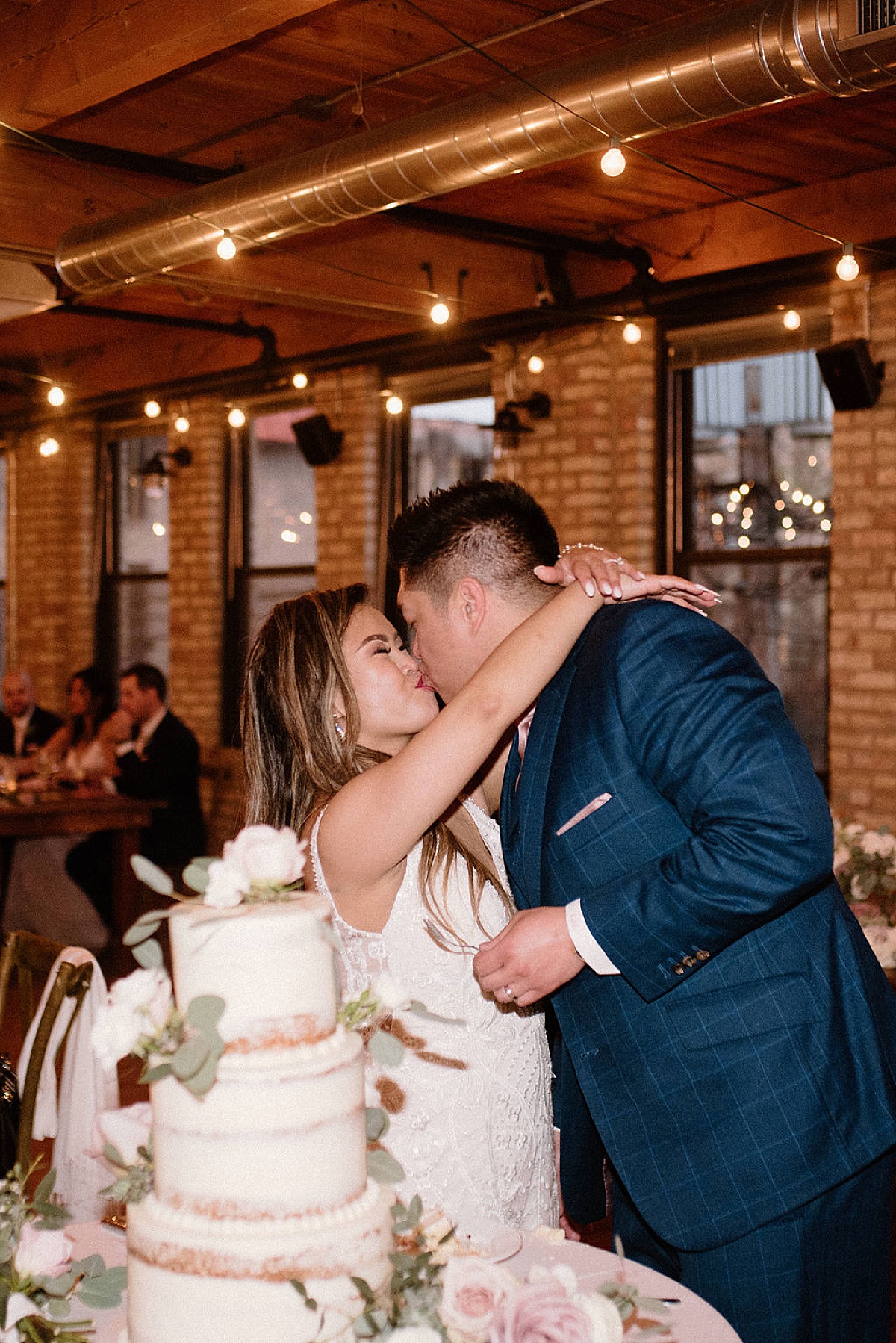 newlywed bride and groom kiss in front of wedding cake during reception shot by Indigo Lace Collective