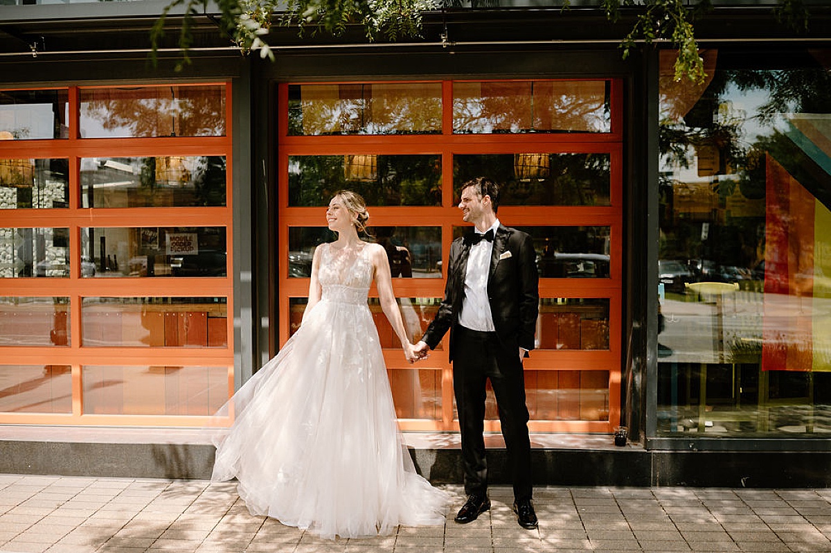 bride in romantic gown and groom hold hands in front of mod cafe before Sami and Andrew's Salvage One Summer Wedding