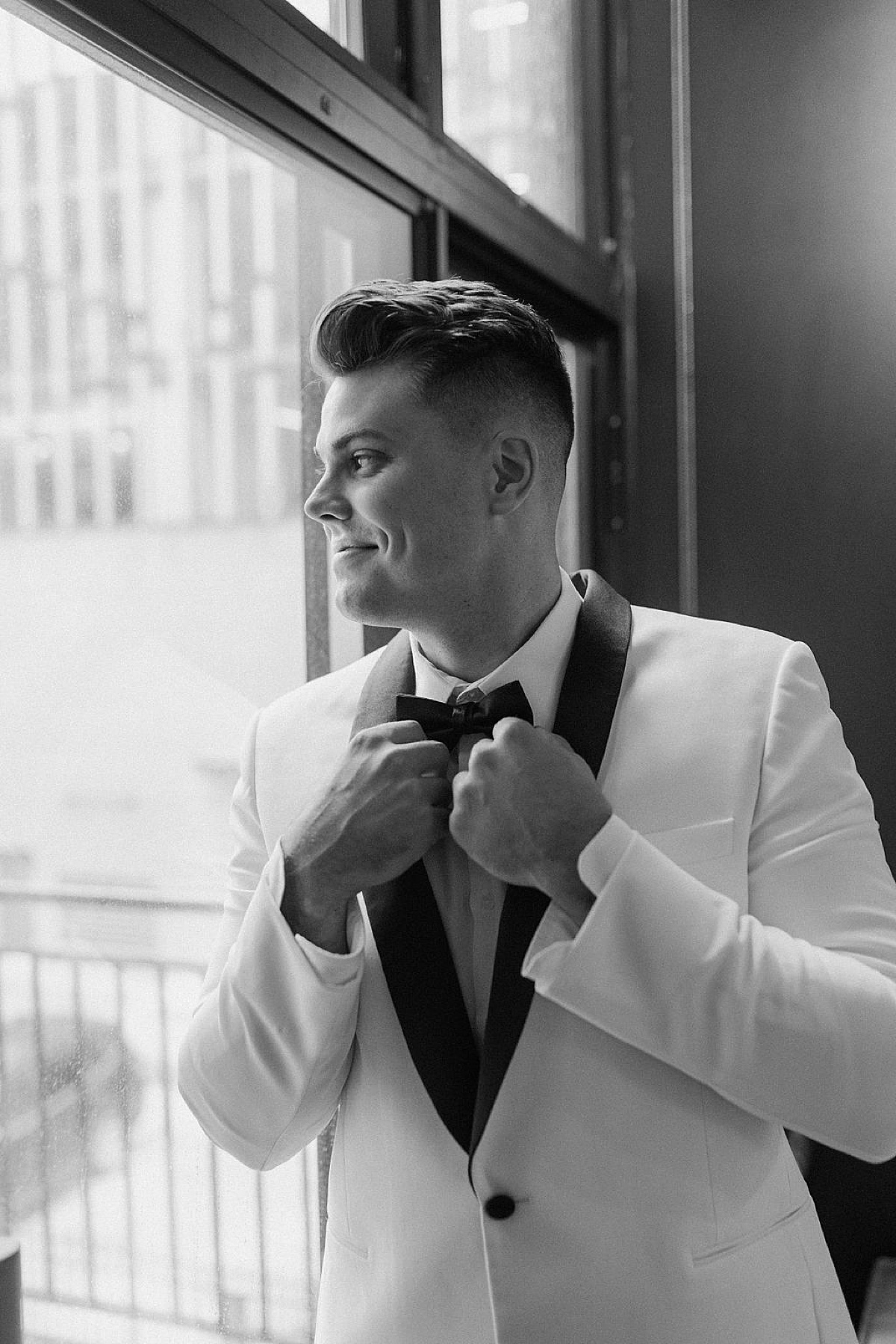 groom in white jacket with black silk lapel looks out window smiling before Glitz & Glam Chicago Wedding