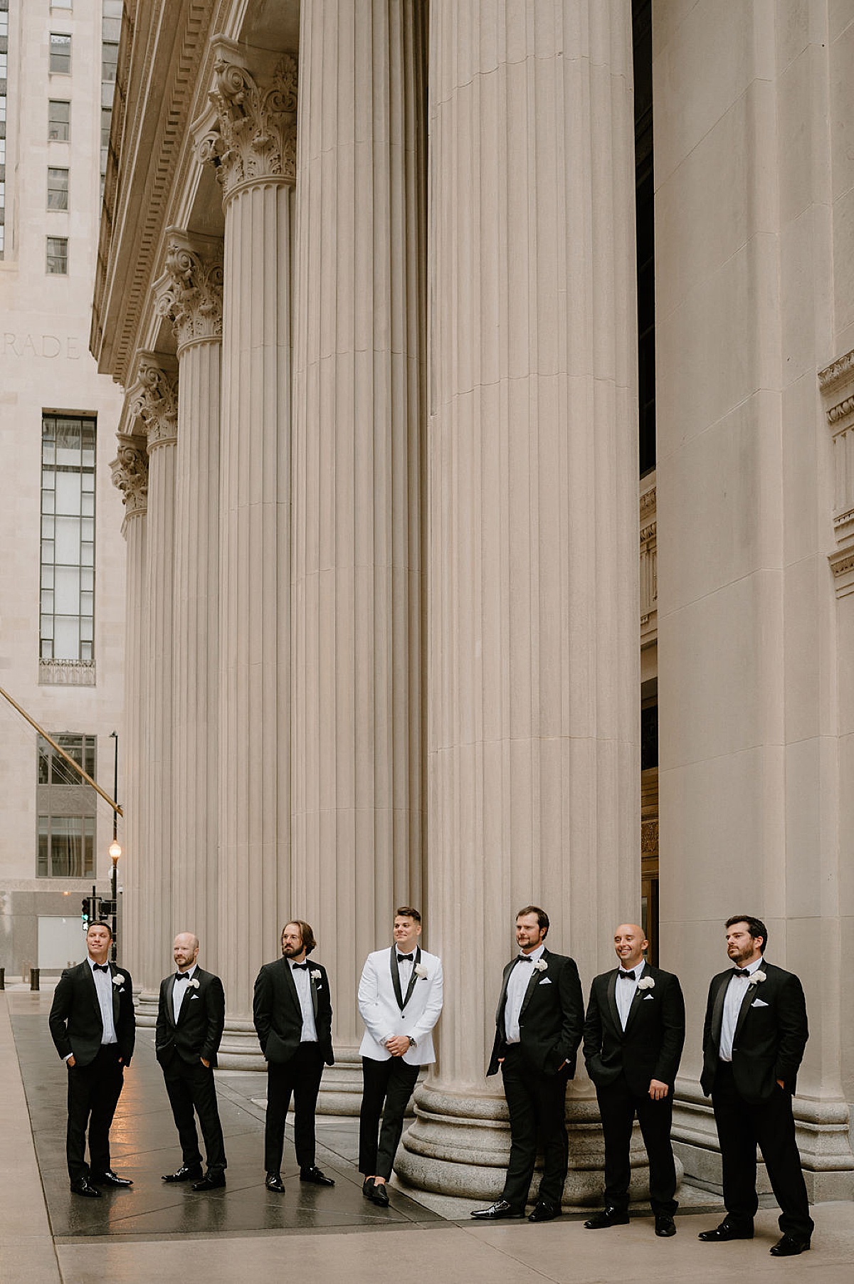 groom in white evening jacket with black lapels poses next to elegant columns with groomsmen shot by Midwest wedding photographer 