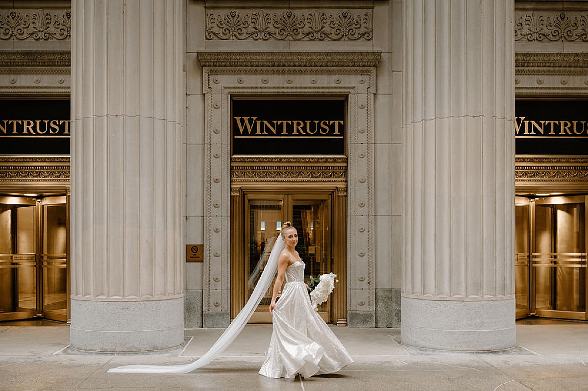 bride with long veil poses in front of gold and stone Chicago building during ceremony shot by Midwest wedding photographer 