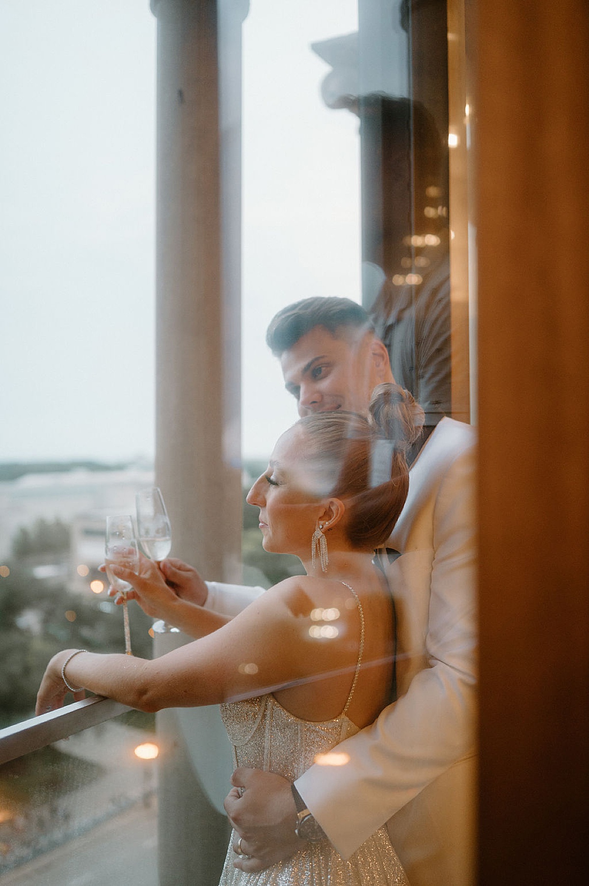 glam bride in glitzy gown and groom pose on balcony with champagne. Indigo Lace Collective