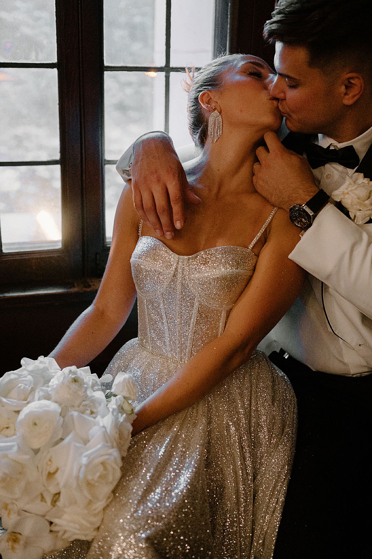 bride in shimmering silver gown holds white rose bouquet and kisses groom in elegant white jacket. shot by Indigo Lace Collective