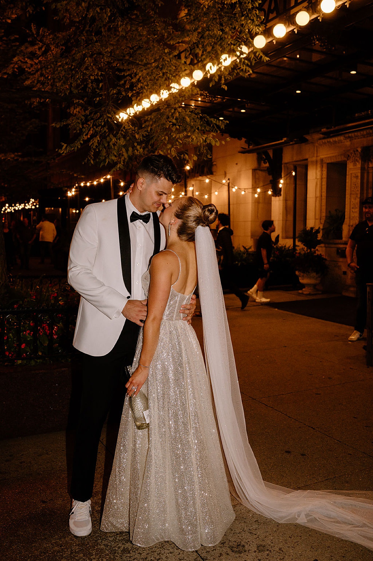 bride with sparkling wedding gown and groom in white jacket kiss over a bottle of champagne after elegant wedding shot by Indigo Lace Collective