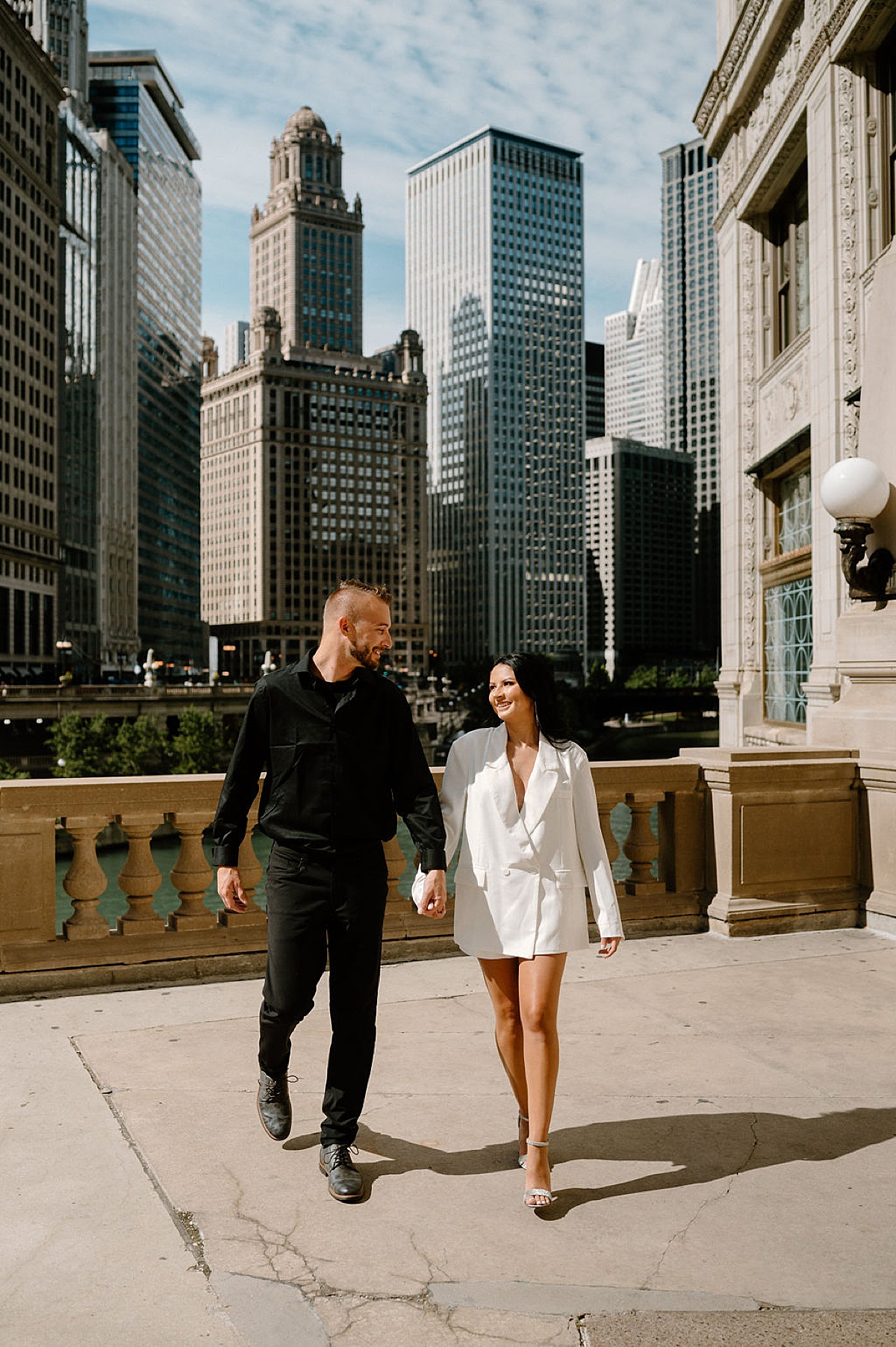 man in black sweater and woman in white blazer walk hand in hand in front of Chicago skyline during big city engagement session