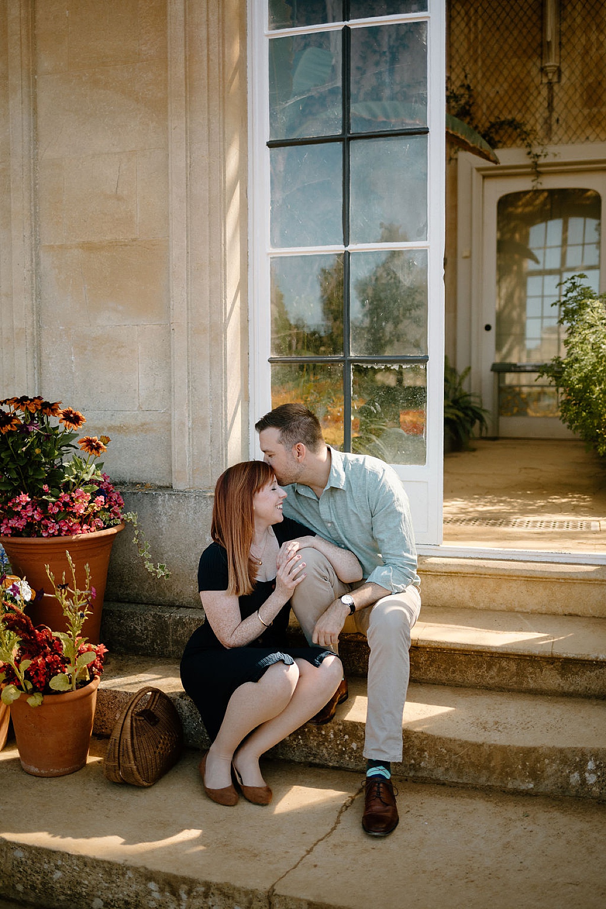 man kisses woman on the forehead sitting on steps of a greenhouse in England during Wrest Park Engagement session