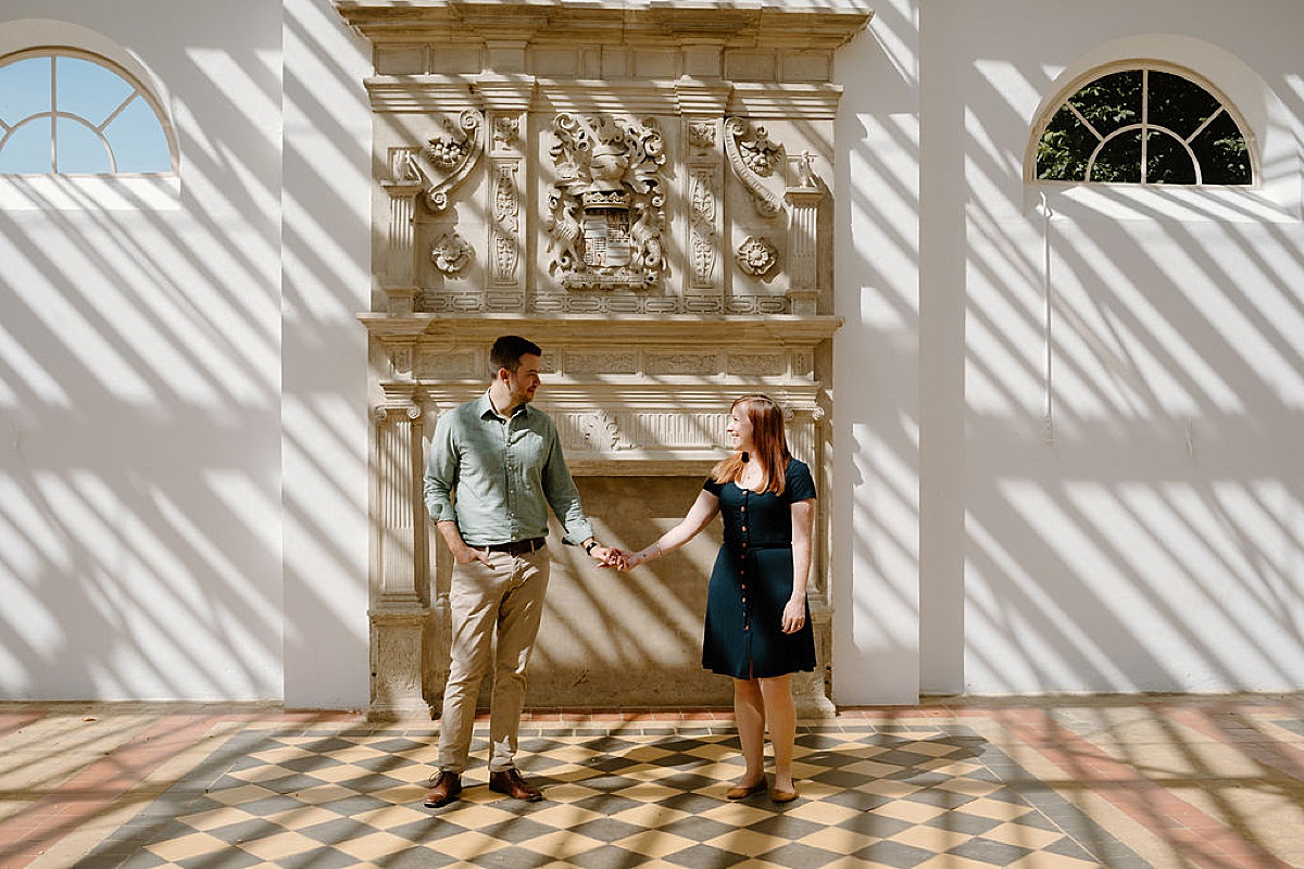 man and woman hold hands and pose in front of antique fireplace at park shot by Indigo Lace Collective