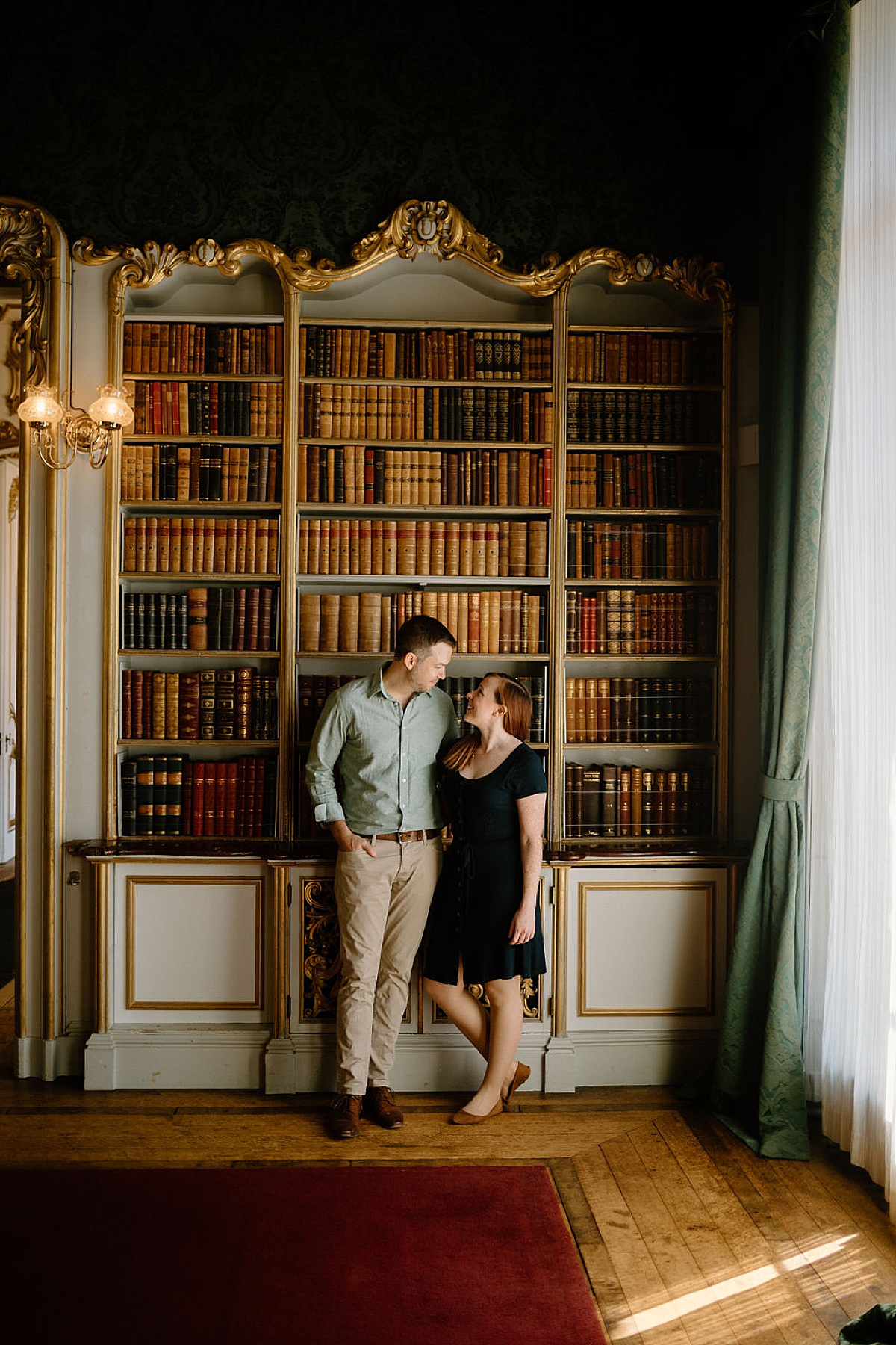 man and woman pose in front of ornate bookshelf during classy engagement shoot by London Wedding Photographer