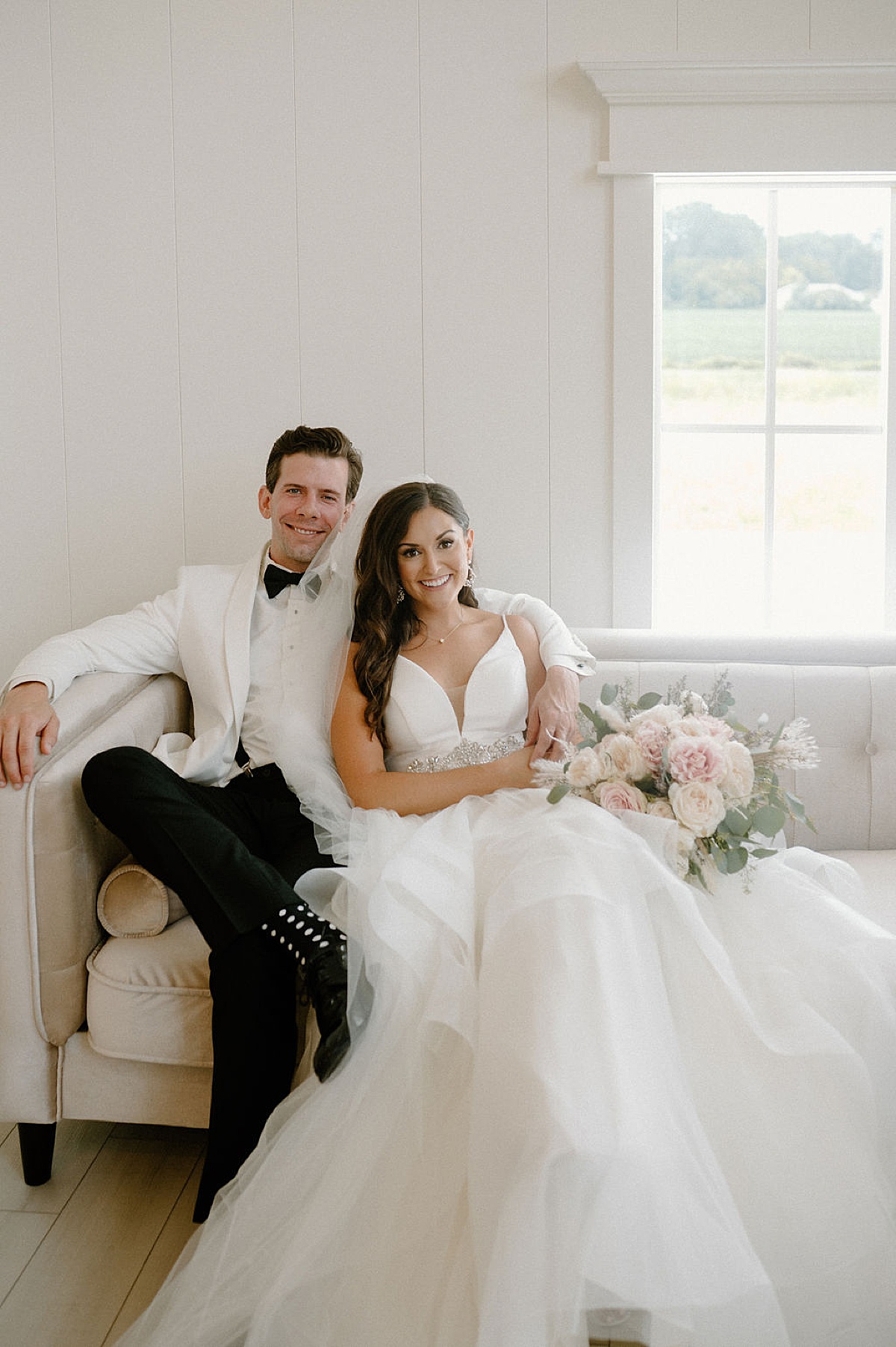 bride in elegant tulle gown with diamond belt sits on white couch with groom in dinner jacket during wedding shot by Indigo Lace Collective