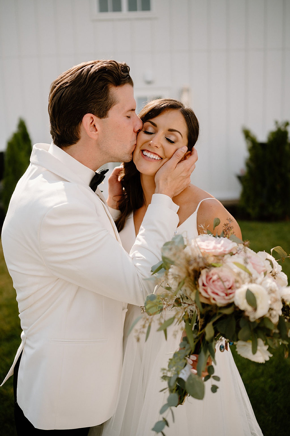 groom in white dinner jacket kisses bride on the cheek as she holds pink and white rose bouquet shot by Indigo Lace Collective