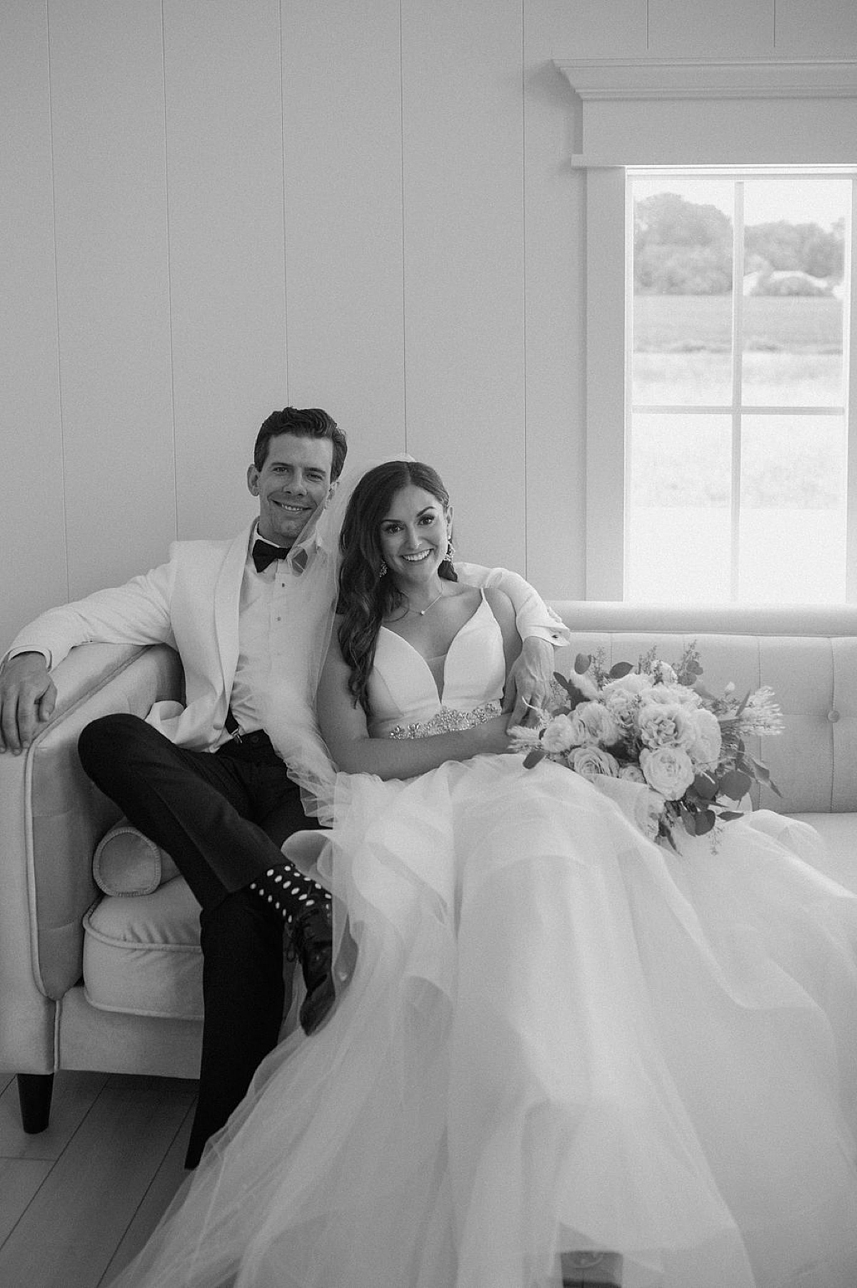 elegant bride in tulle gown and dapper groom in white coat pose on leather couch before ceremony shot by Michigan wedding photographer