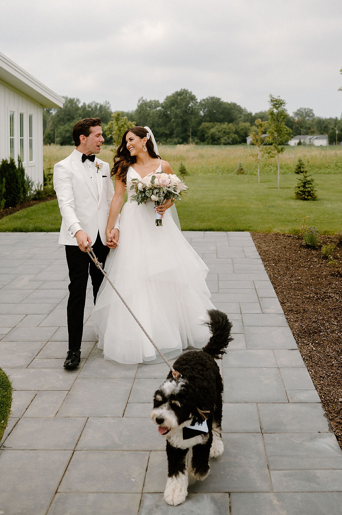 Bride in tulle gown walks hand in hand with groom in white dinner jacket as they walk doodle puppy on a leash at ceremony shot by Michigan wedding photographer
