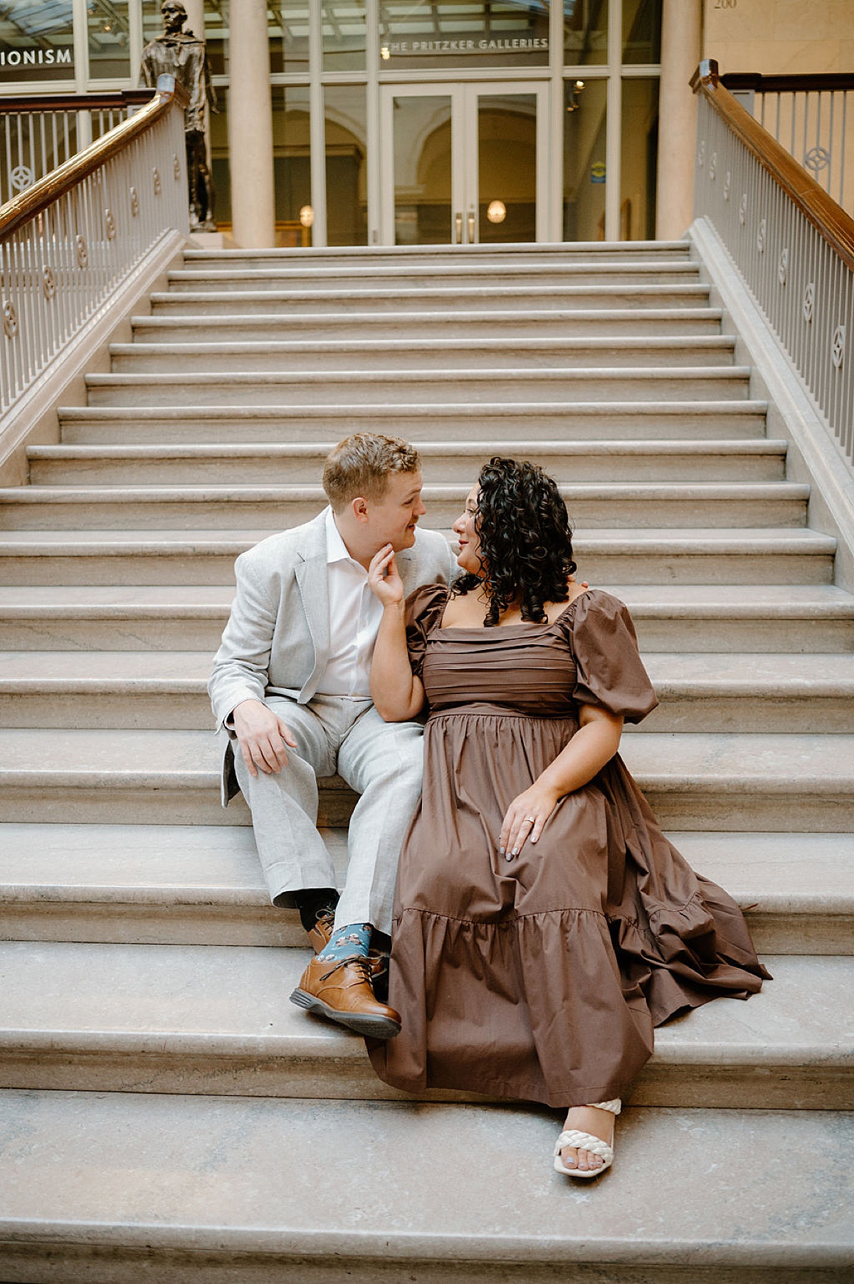 man in gray suit and woman in romantic dress sit on art museum steps during shoot by Indigo Lace Collective