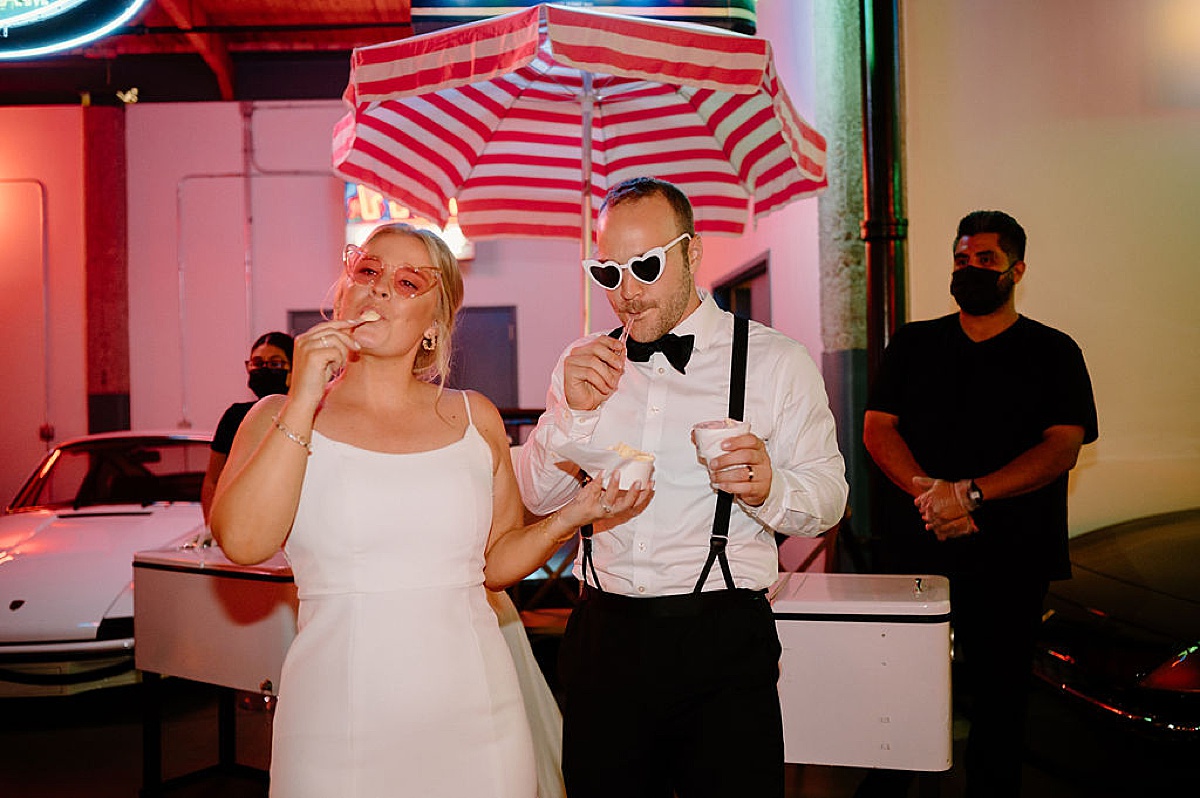 bride and groom in fun sunglasses eat ice cream at reception at vintage car warehouse shot by midwest wedding photographer