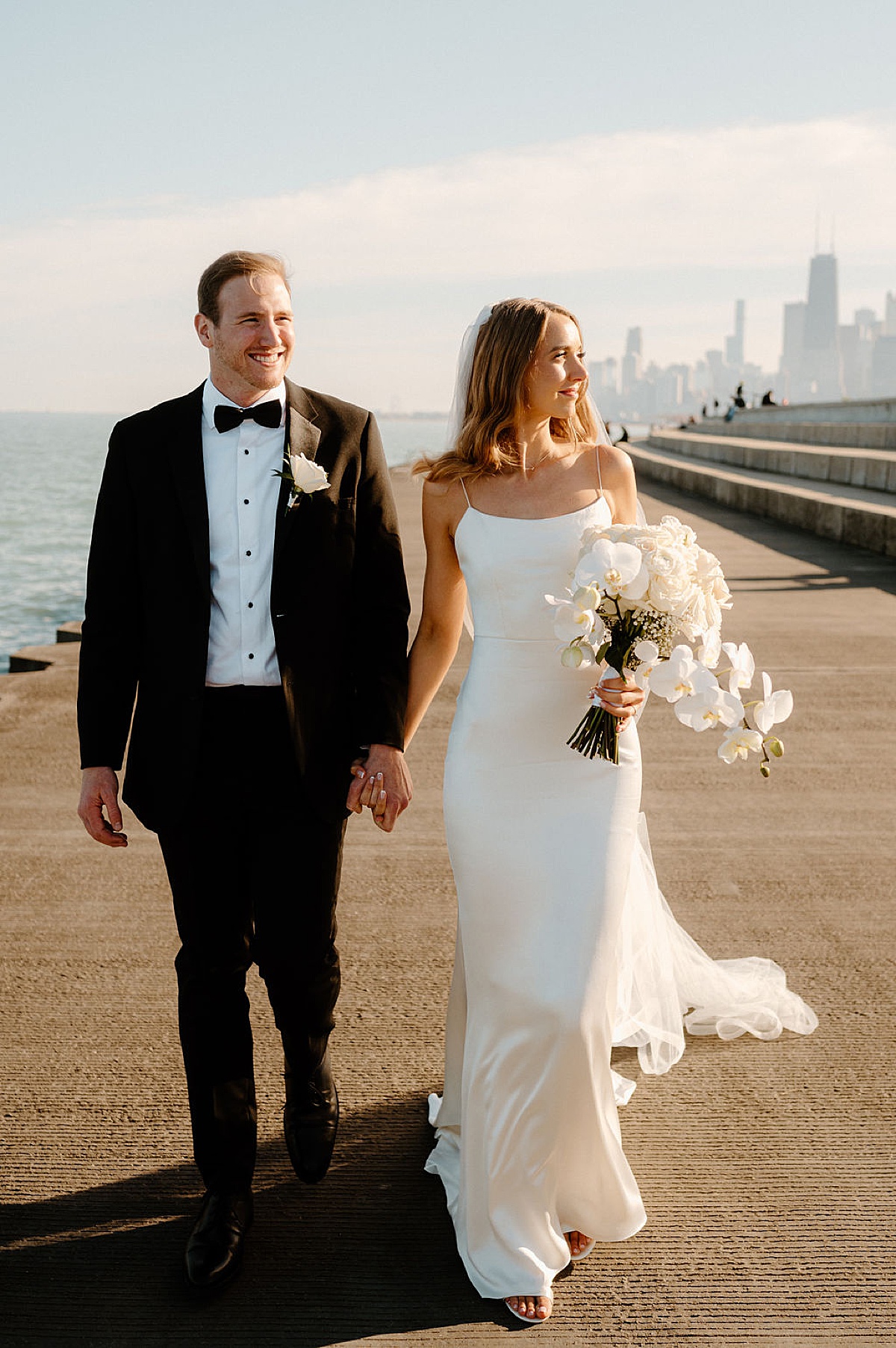 bride with white orchid and rose bouquet walks with groom on the waterfront in chicago before Indigo Lace Collective