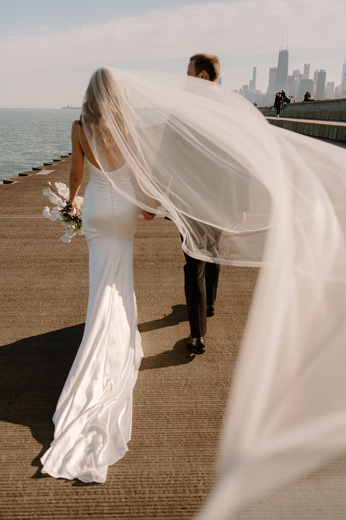 bride in elegant gown and white orchid bouquet walks city waterfront with groom before wedding shot by Indigo Lace Collective