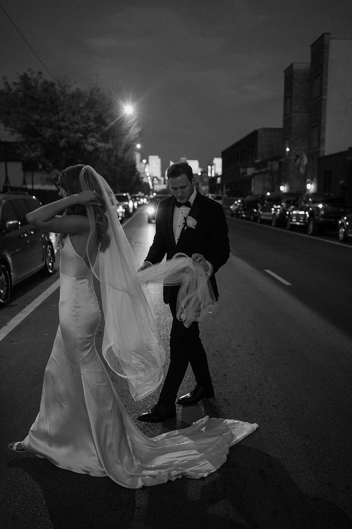 bride in silk gown and long veil crosses the street with groom during noir shot by Chicago wedding photographer