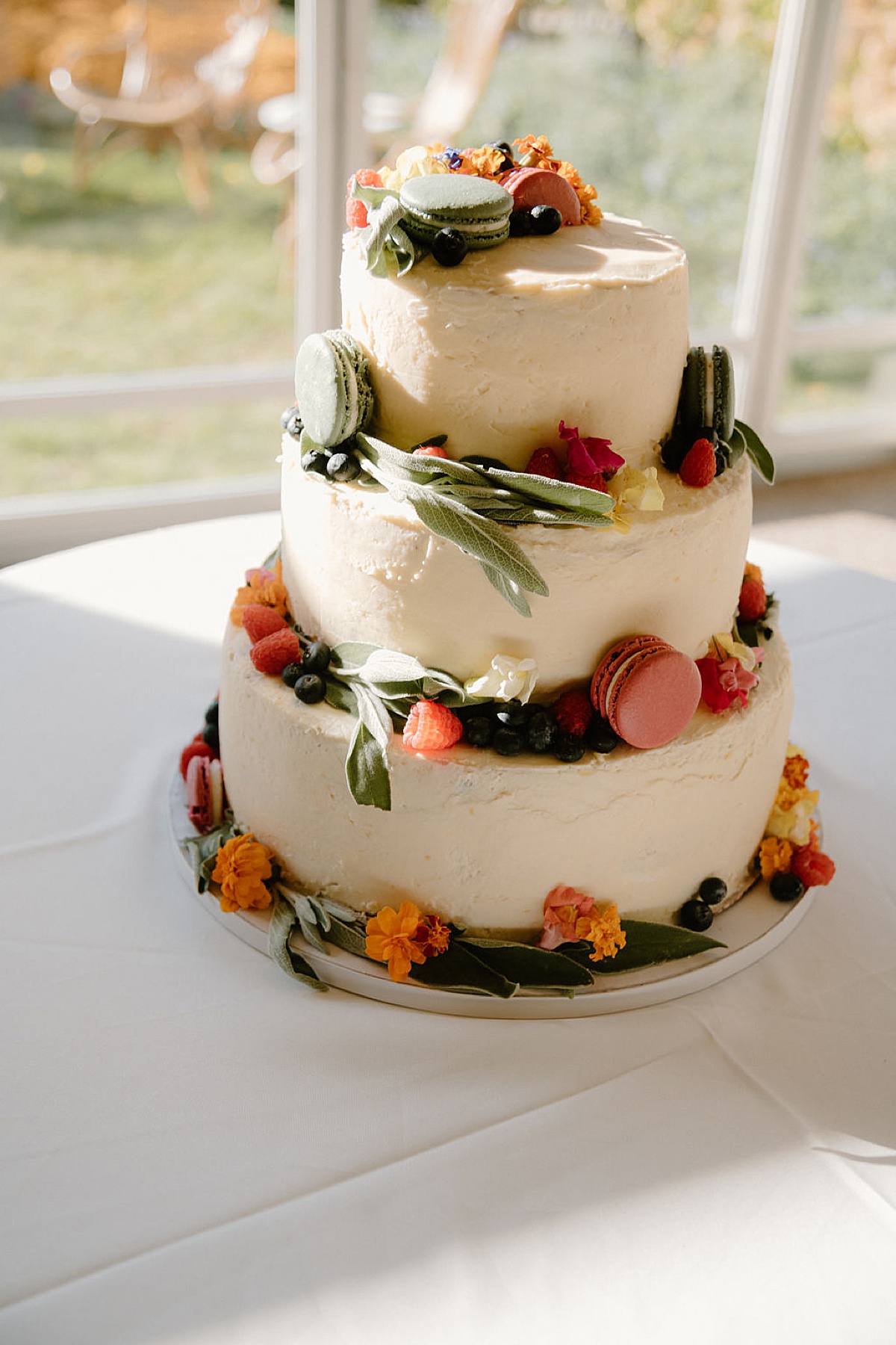 wedding cake with sage leaves, macarons, and edible flowers waits for bride and groom at reception shot by Indigo Lace Collective