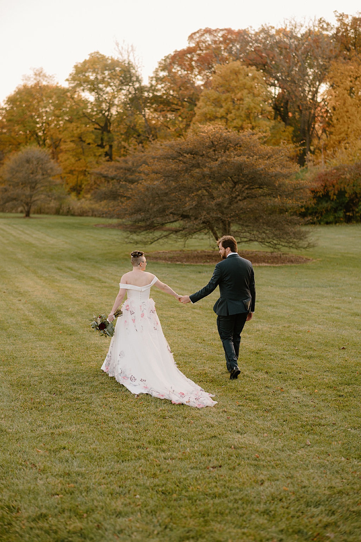 bride in romantic trailing gown and groom hold hands and walk across a field shot by midwest wedding photographer