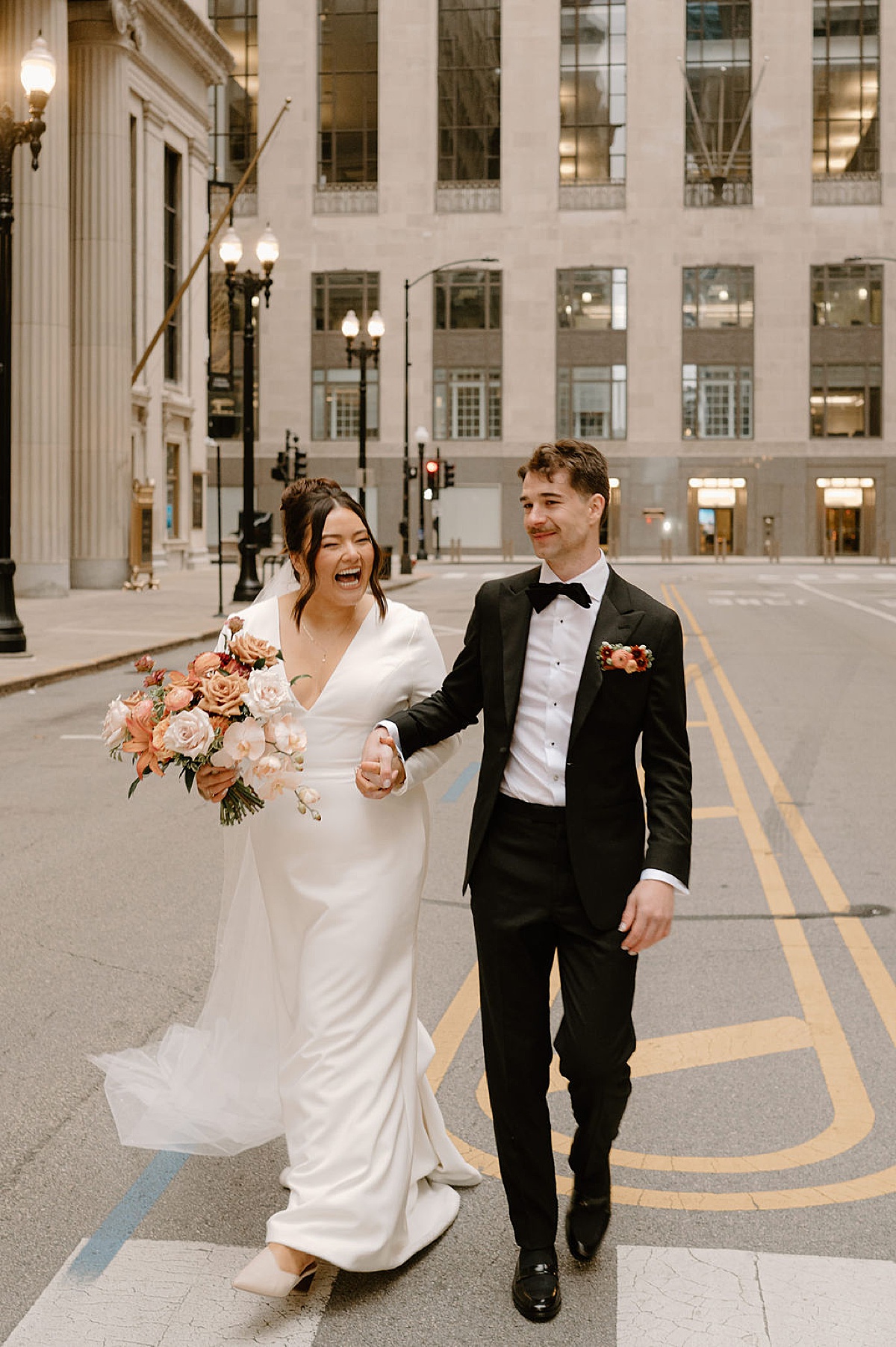 elegant bride in minimalist gown poses with groom and bouquet of peach and rust colored flowers shot by Chicago wedding photographer