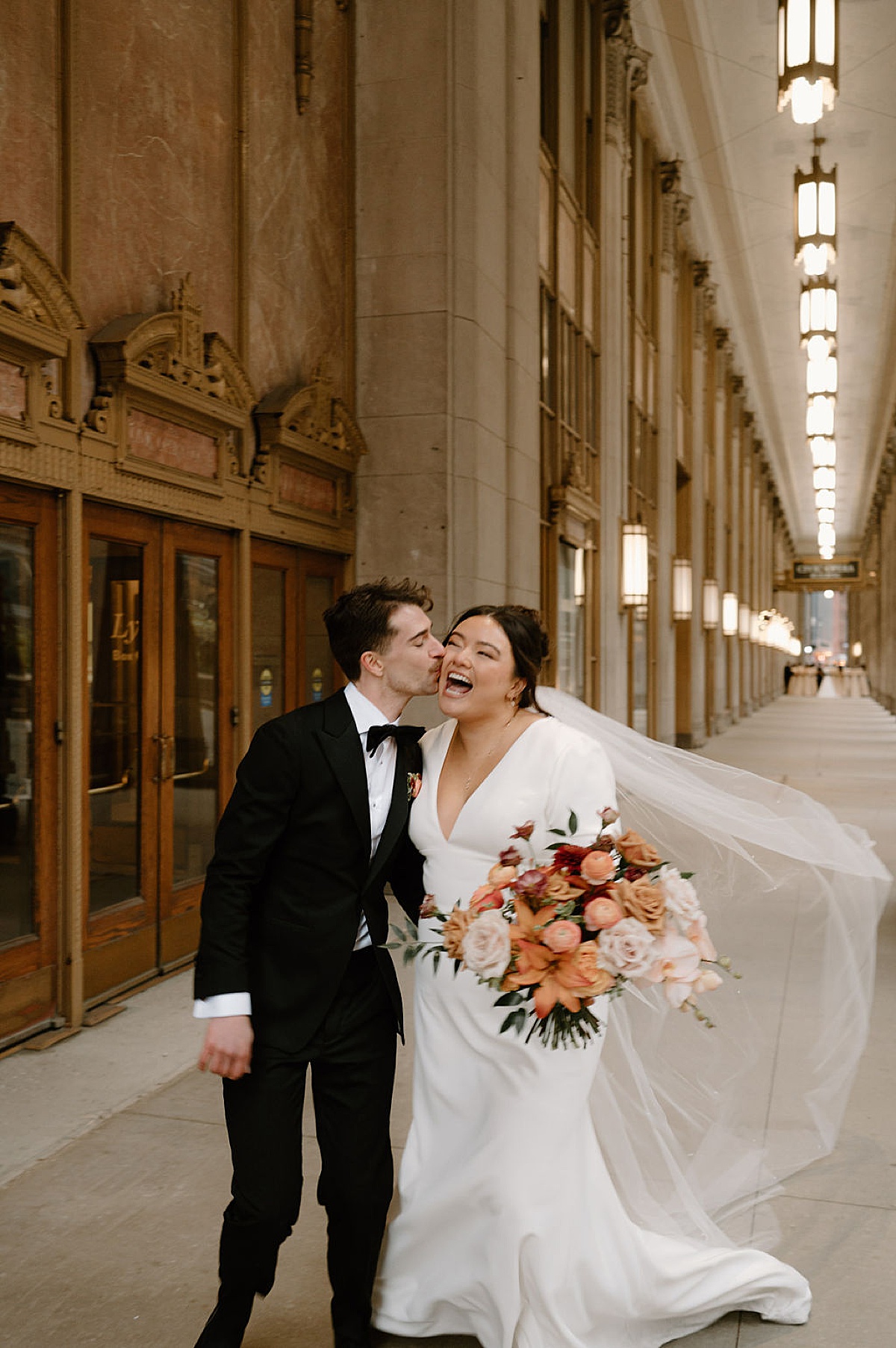 groom steals a kiss on the cheek from laughing bride holding autumnal bouquet shot by Indigo Lace Collective
