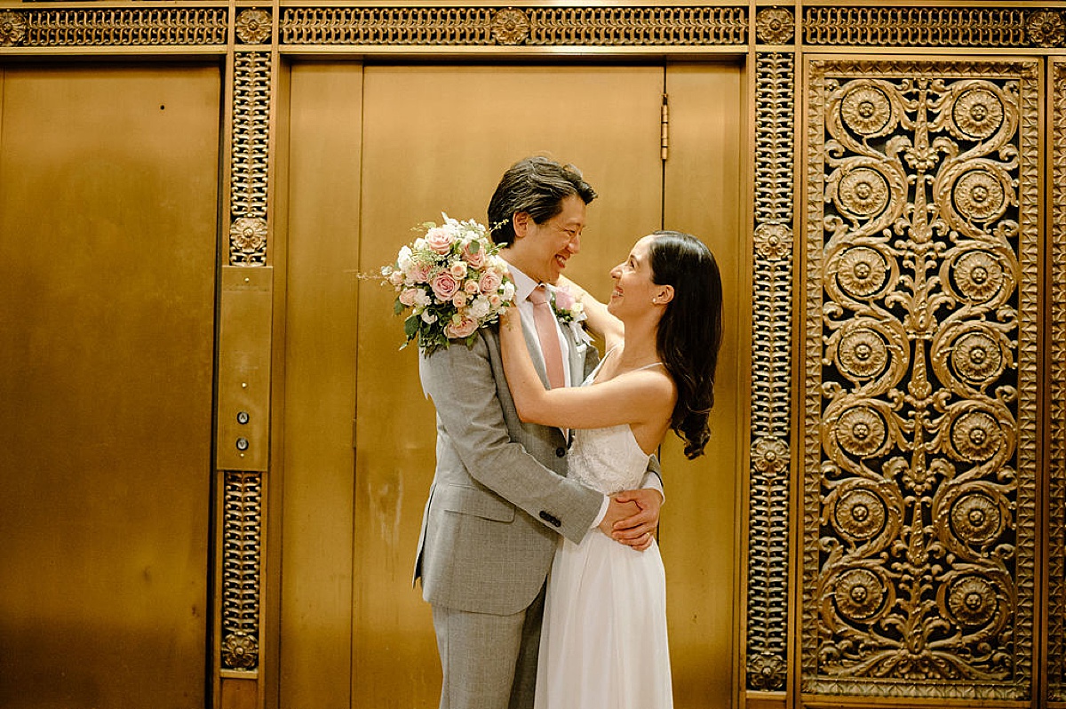 groom in dapper grey suit and bride in delicate gown embrace in front of ornate gold doors during city hall sweetheart elopement