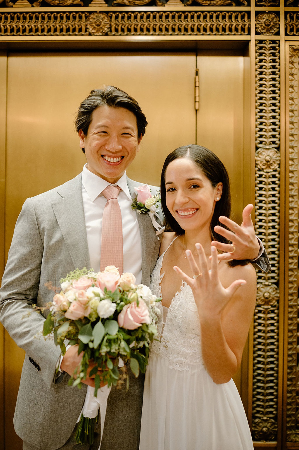 groom in dapper gray suit and bride in delicate gown show off wedding rings in front of gold doors during city hall sweetheart elopement