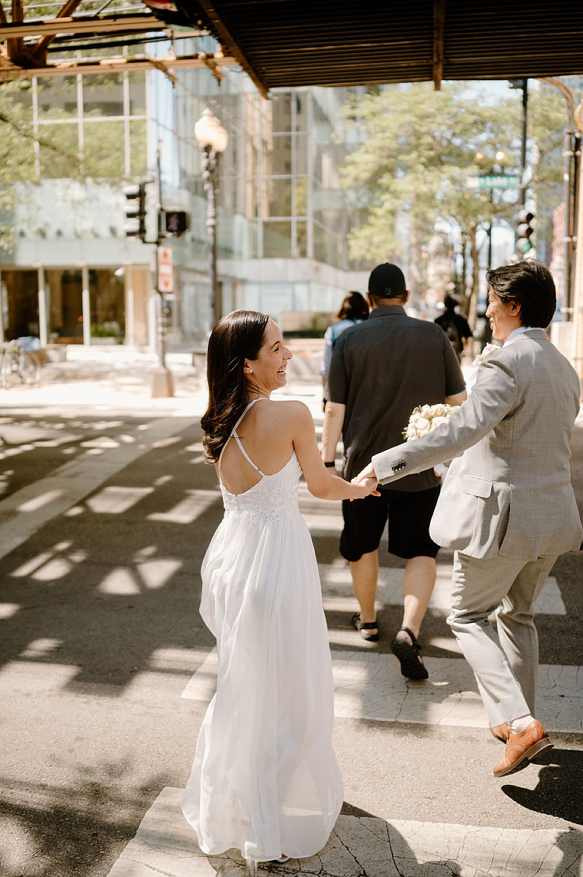 groom grabs bride's hand as they laugh and cross the street under Chicago's L train after city hall sweetheart elopement
