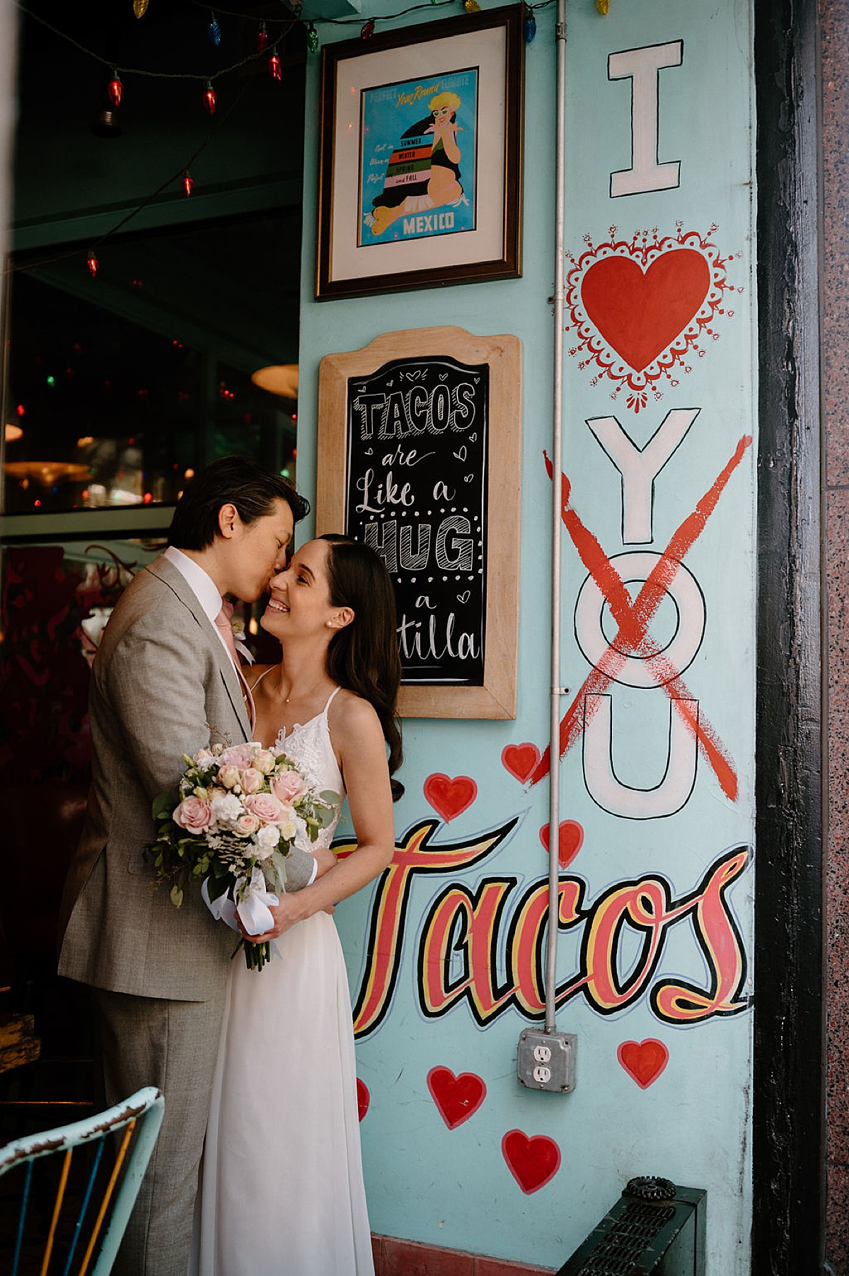 groom in gray suit and pink tie kisses bride in delicate gown on the cheek at taco shop | Chicago wedding photographer