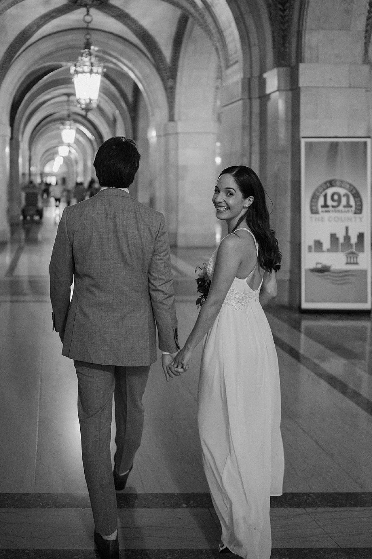 bride in delicate gown looks over her shoulder smiling as she walks with groom through marble hall shot by Indigo Lace Collective