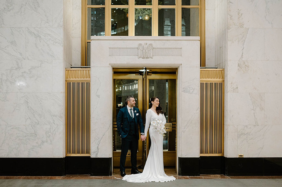 bride in elegant gown with train poses with groom at gold doors before celebration at old Chicago post office