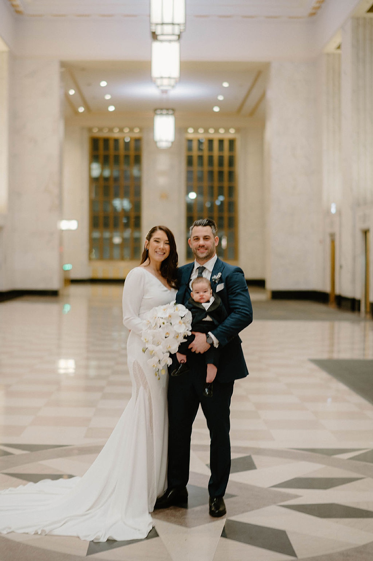 bride and groom hold infant son in elegant marble hallway during post-covid celebration shot by Chicago wedding photographer