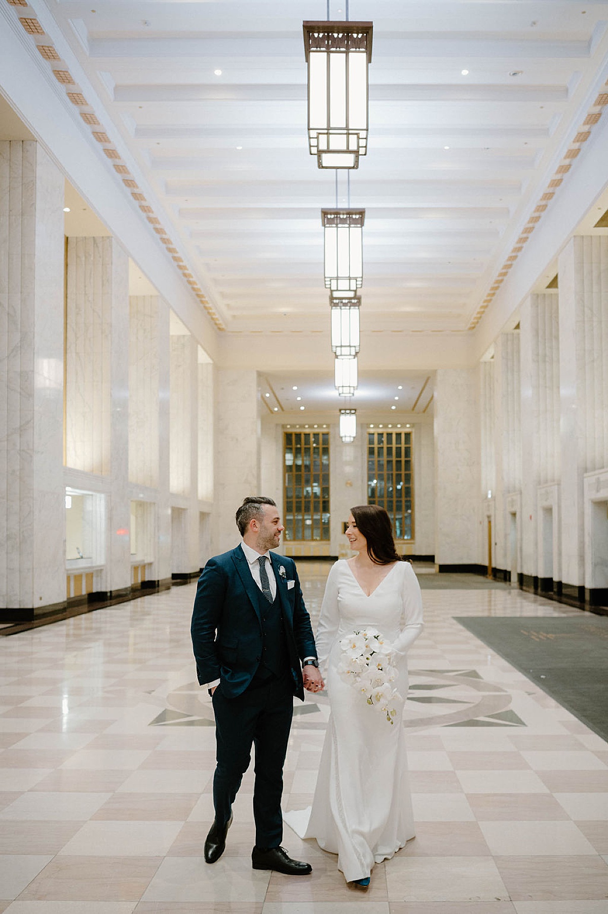 bride in simple gown and orchid bouquet poses with groom in marble hall after wedding shot by Indigo Lace Collective